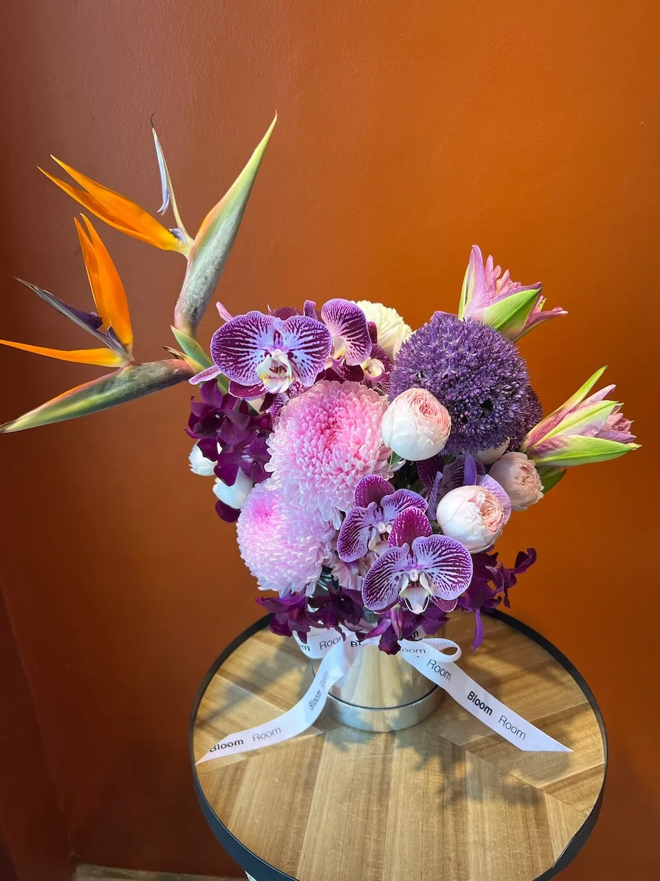 Striking arrangement with striped phalaenopsis orchids, birds of paradise, purple allium and blush peonies in a silver vase
