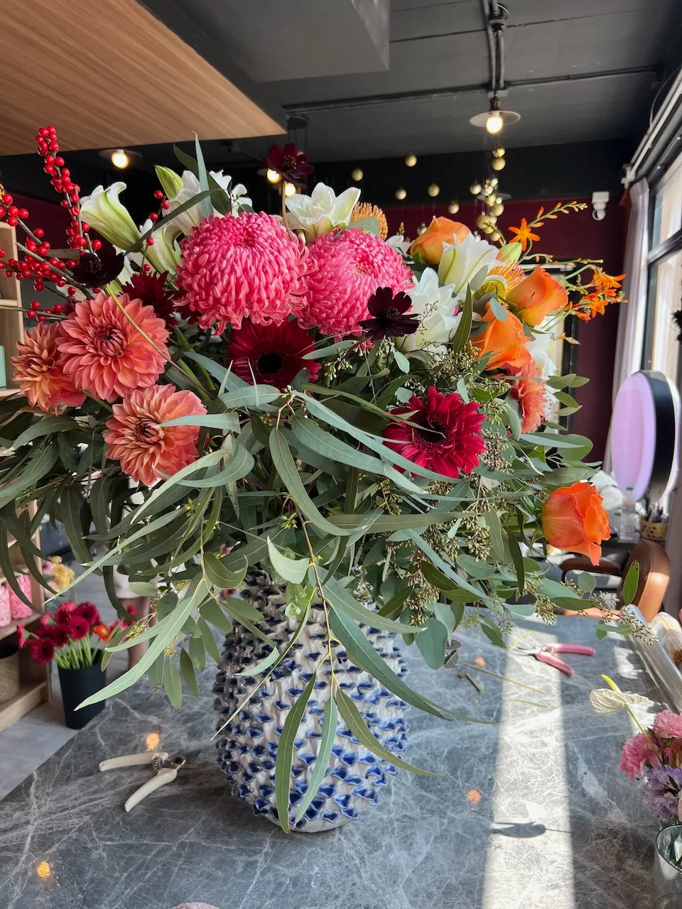 Large arrangement of hot pink chrysanthemums, coral dahlias, red ilex berries and white lilies in a blue-and-white ceramic vase