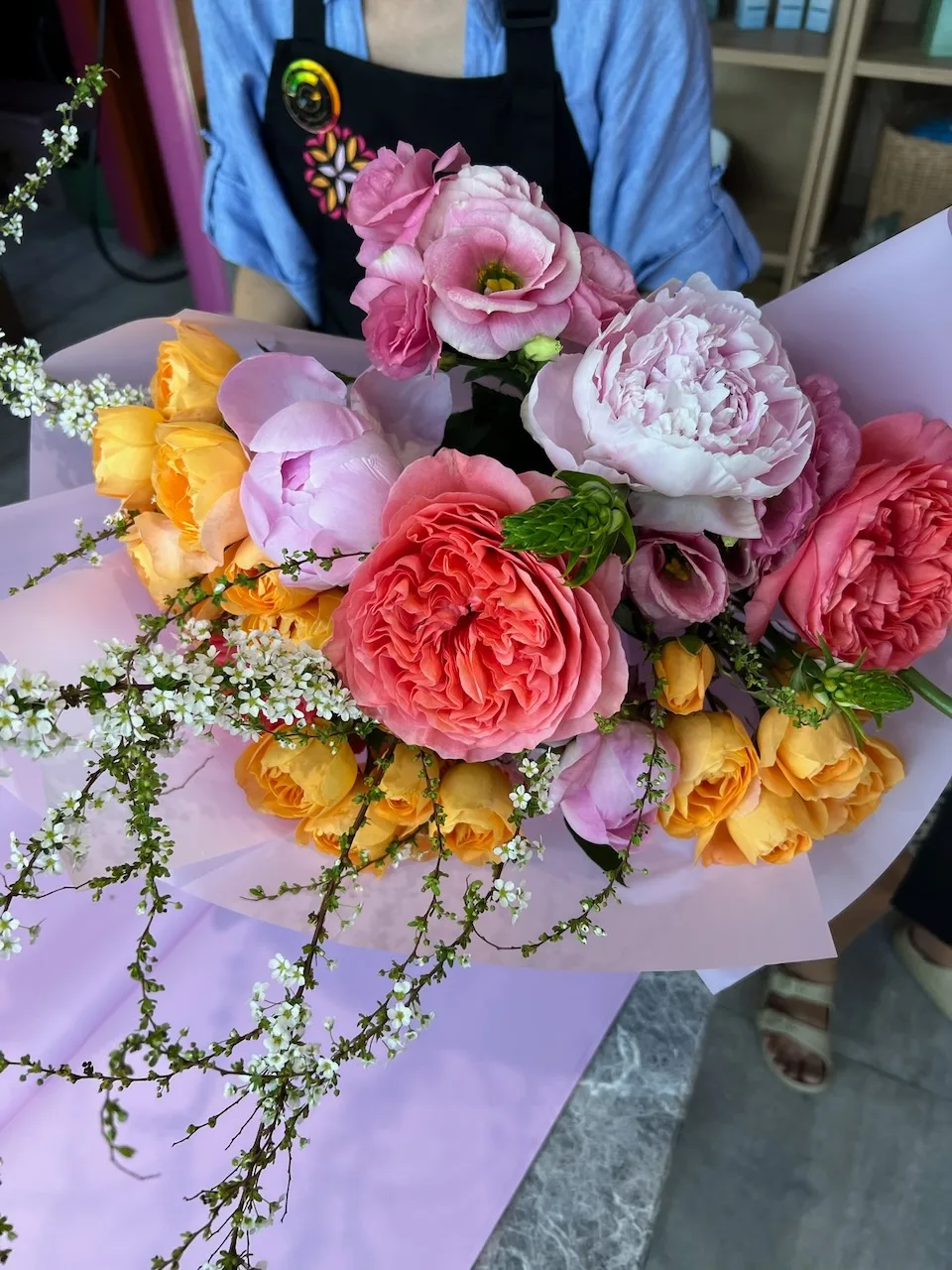 Vibrant hand-tied bouquet with coral peonies, yellow roses and pink lisianthus wrapped in lavender paper