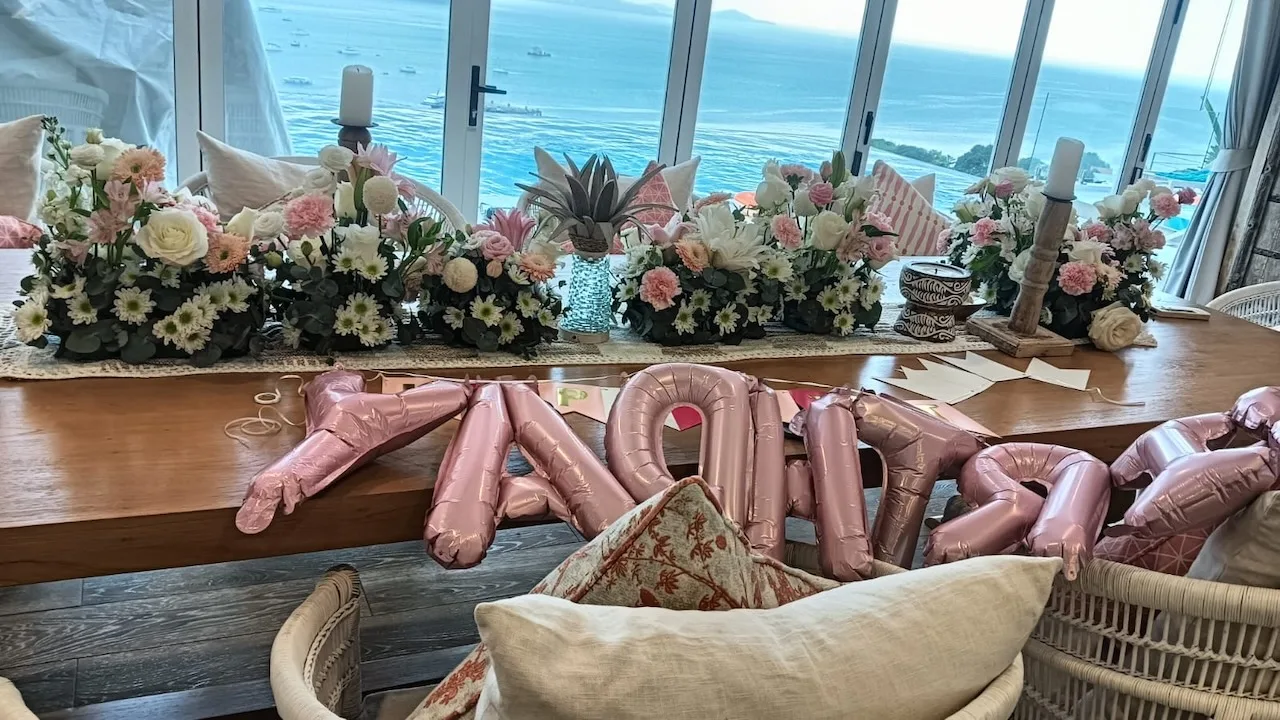 Pastel pink and white arrangements line a table with balloon letters against a sea view