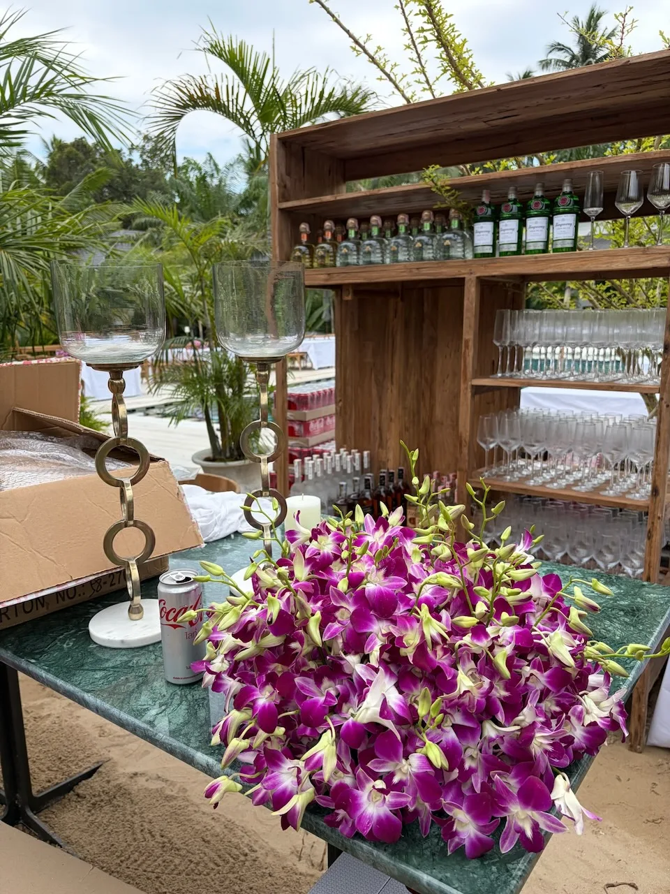 Pile of purple dendrobium orchids on green marble bar with gin bottles and glassware at poolside event setup