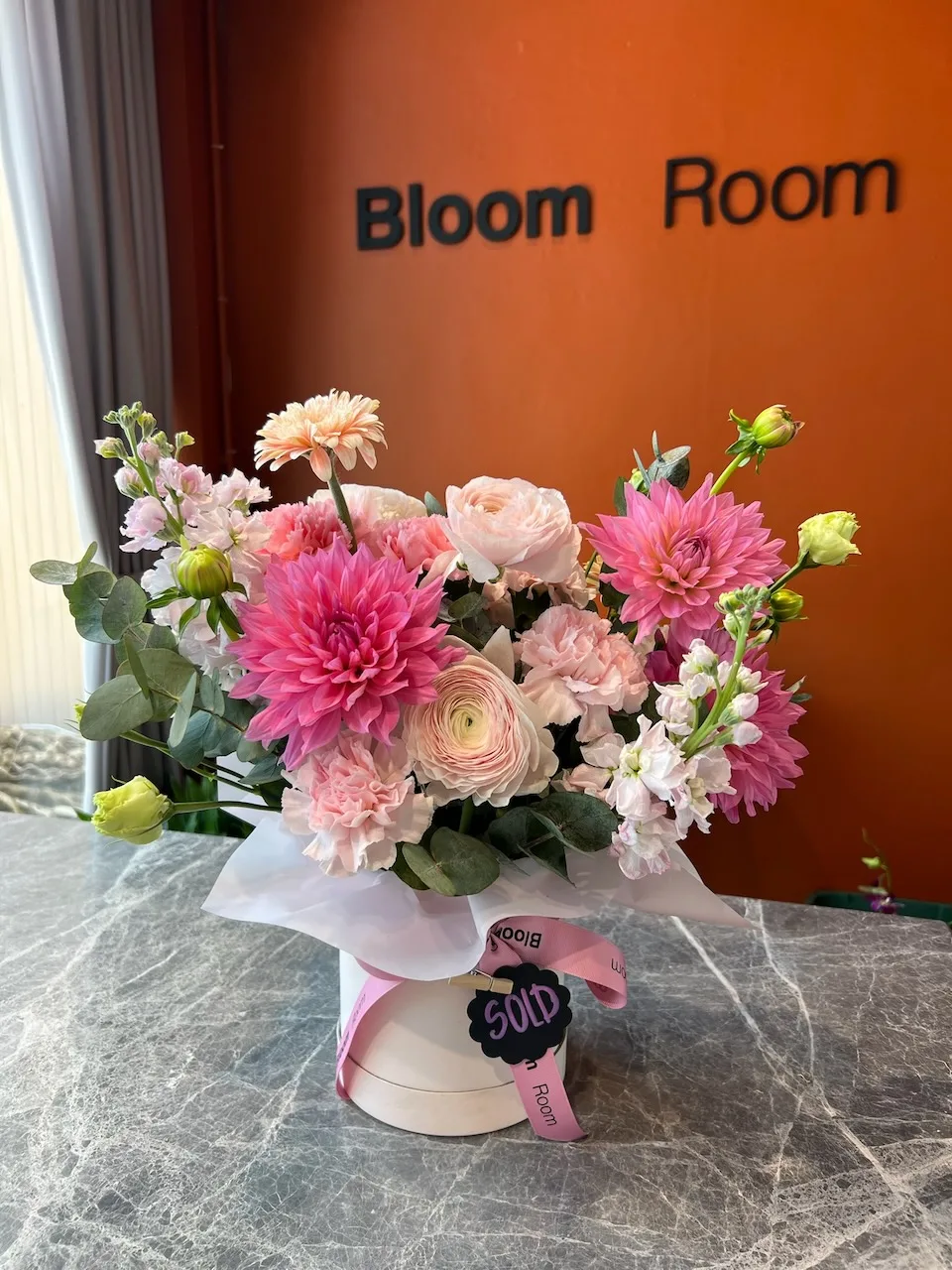 Шляпная коробка с розовыми георгинами, ранункулюсами и хризантемами на мраморе под вывеской Bloom Room