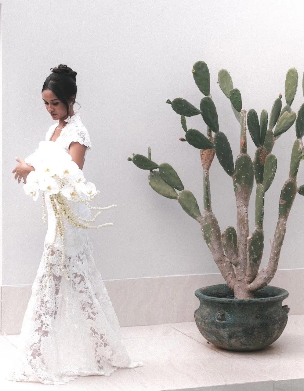Bride in white lace gown with dramatic cascading orchid bouquet beside a cactus